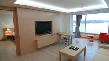 Una moderna camera d'albergo con una TV a schermo piatto fissata a un pannello di legno, un tavolino con un telecomando, una poltrona rossa e un'ampia finestra con vista su un corpo d'acqua.