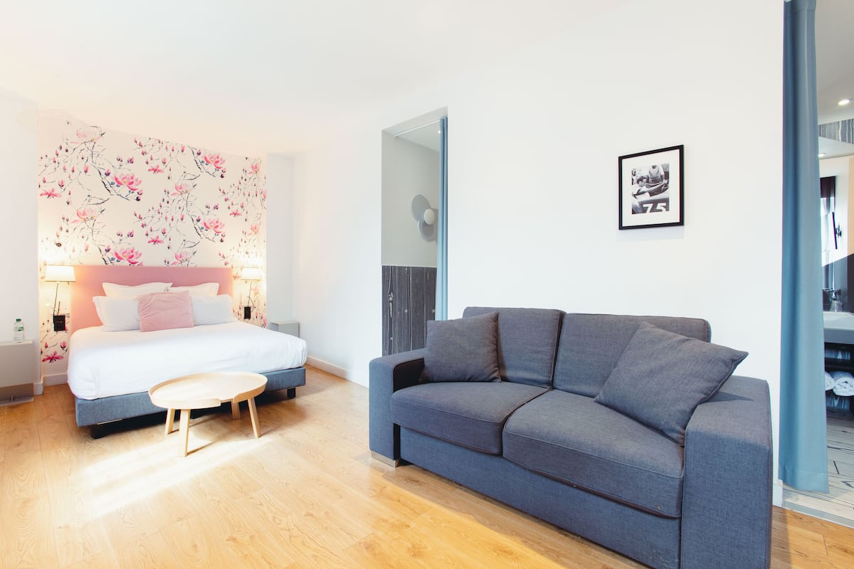 A modern bedroom with a bed, bedside lamps, a grey sofa, and a wooden floor.