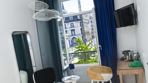 Quarto com vista para a cidade, uma pequena mesa com uma cadeira, uma TV fixada na parede e uma janela com cortinas.