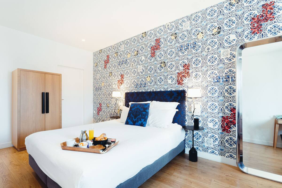 A neatly made bed with a tray of breakfast items, a wooden wardrobe, and a patterned wall.