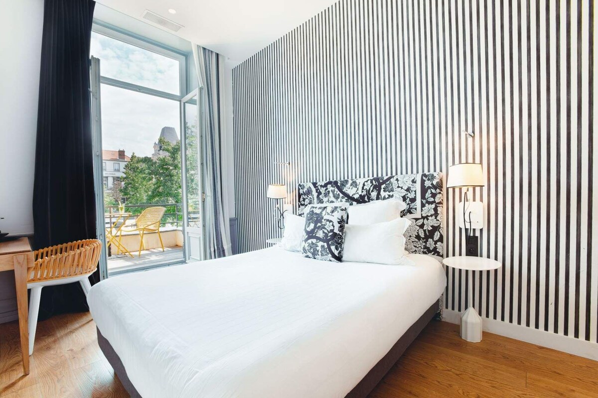 A modern hotel room with a large bed, a desk, and a balcony with yellow chairs.