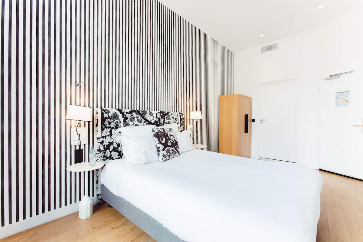 A modern hotel room with a large bed, a wooden wardrobe, a bedside table with a lamp, and a wall with vertical stripes.
