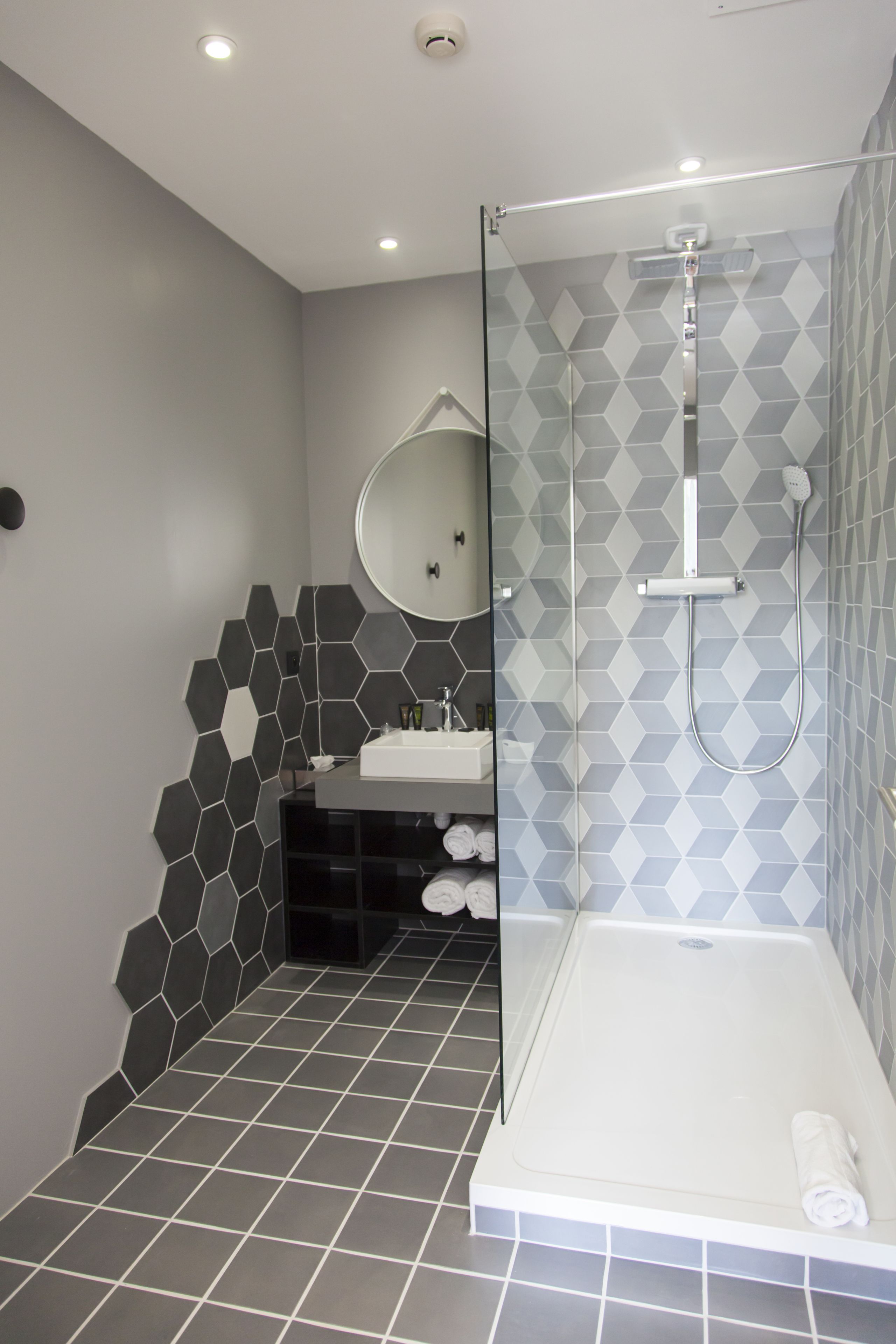 Une salle de bain moderne dotée d’une douche à parois de verre, d’un miroir rond et d’un lavabo avec un pare-éclaboussures en carreaux hexagonaux.