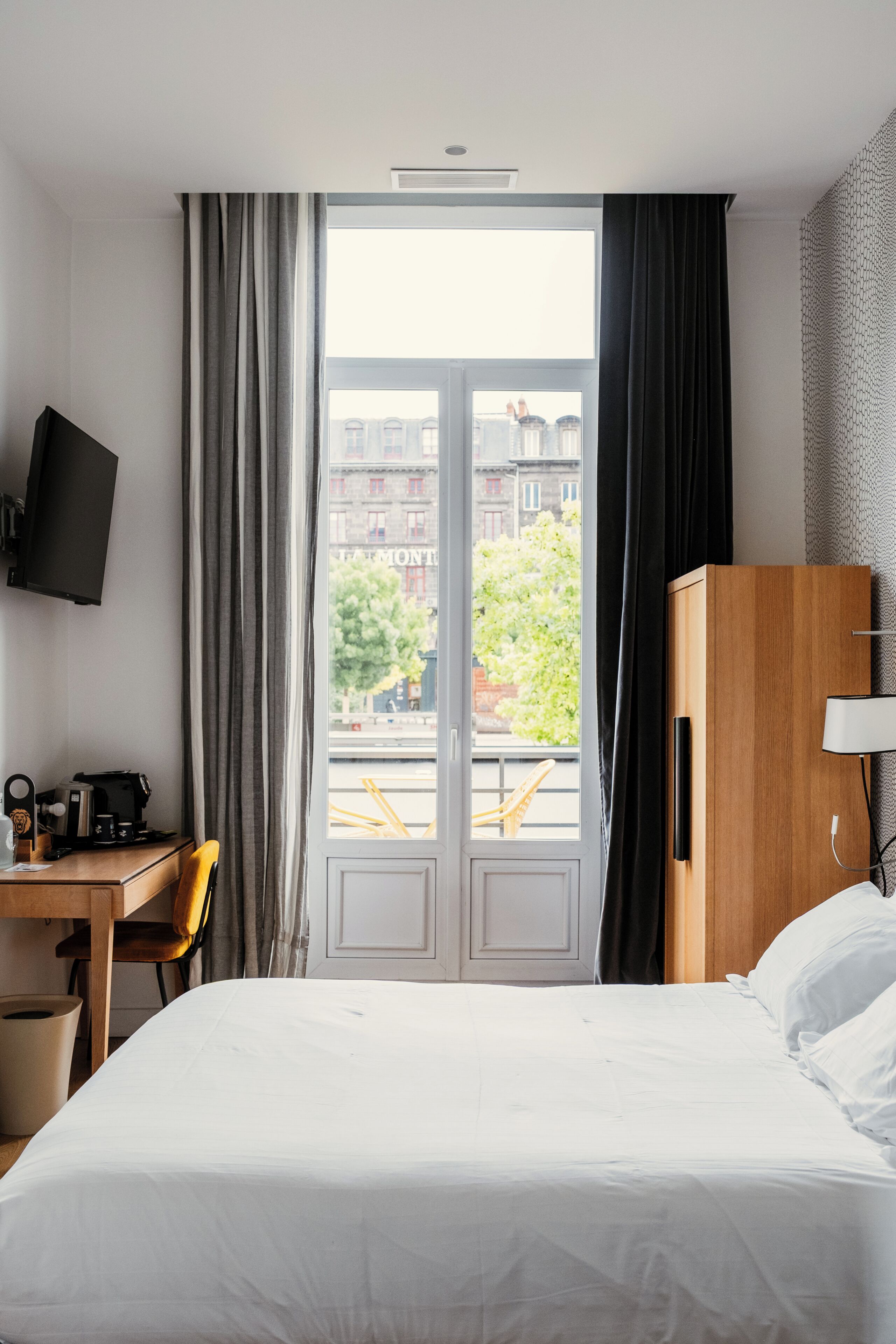 Habitación de hotel con cama, escritorio con silla, televisión y vista a la ciudad a través de un ventanal grande.
