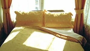 Un lit avec une tête de lit à motifs et des oreillers, des rideaux et la lumière du soleil qui entre par une fenêtre.
