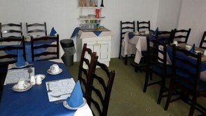 Un espace repas avec des tables dressées pour quatre personnes, chacune étant recouverte d’une nappe bleue et agrémentée d’assiettes blanches. Des chaises en bois sont disposées autour des tables, et un buffet blanc présente divers objets sur son dessus.