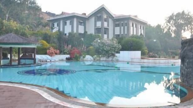 A swimming pool with a clear blue center, surrounded by a paved area and a building with a classical architectural style in the background. - Blue Country Resort (Mahabaleshwar)