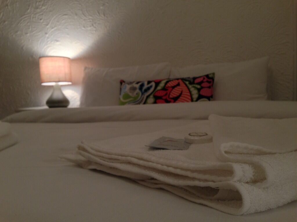 Una cama bien hecha con una toalla blanca doblada, una botella pequeña, un cojín colorido y una lámpara sobre una mesita de noche.