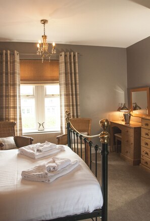 A bedroom with a bed, a dresser, a chair, a window with curtains, and a chandelier.