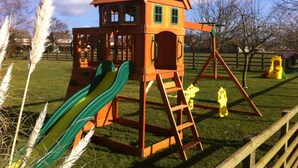 A wooden playhouse with a slide and ladder, surrounded by grass and trees. - The Poplars (Thirsk)