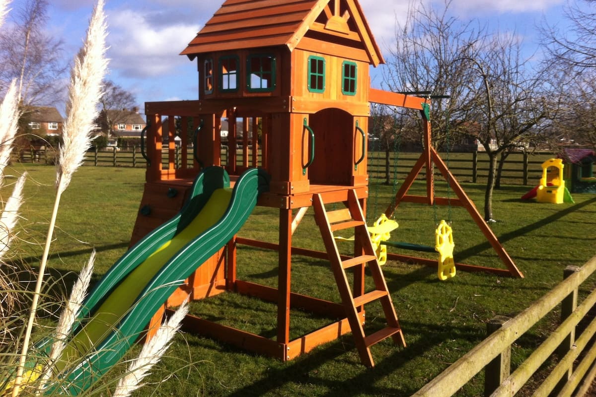 A wooden playhouse with a slide and ladder, surrounded by grass and trees.
