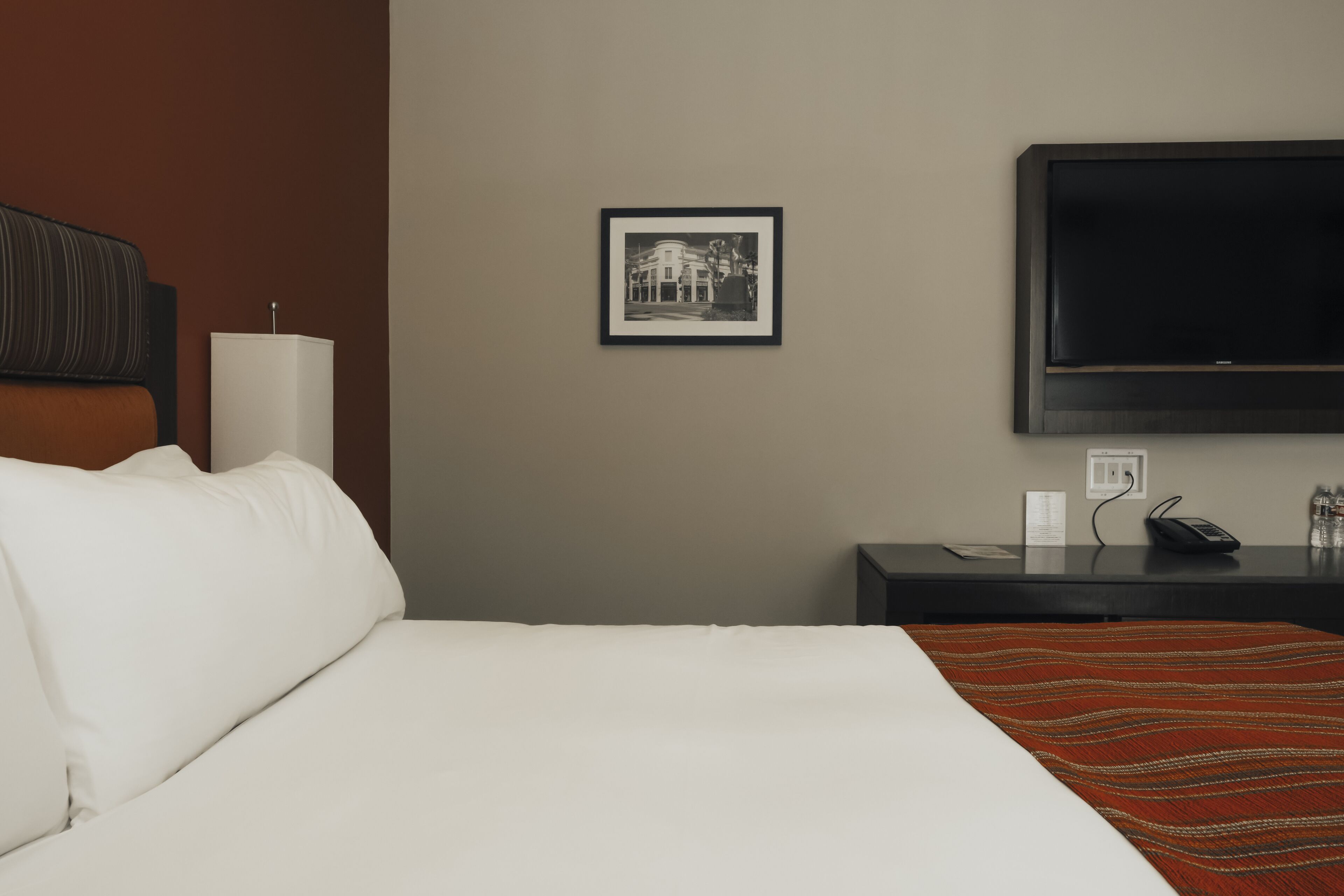 A hotel room with a bed, a TV mounted on the wall, a framed picture, a bedside table, and a lamp.