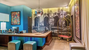 A hotel lobby with a reception desk, two blue cushioned seating areas, a decorative cart, and a mural of a garden scene. - SIRTAJ - Beverly Hills (Beverly Hills)