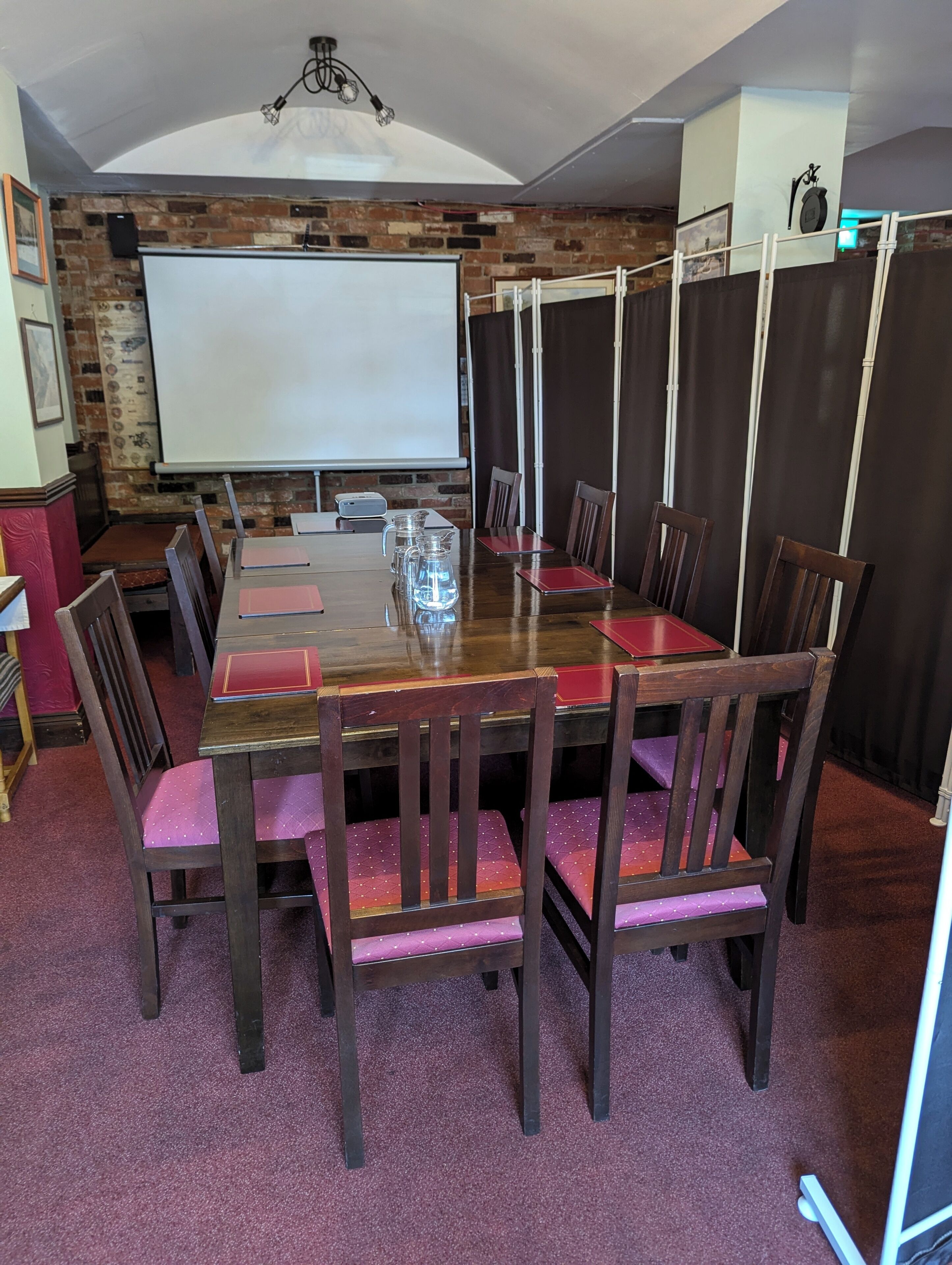 A conference room with a long wooden table, red cushioned chairs, a whiteboard, and a projector screen.