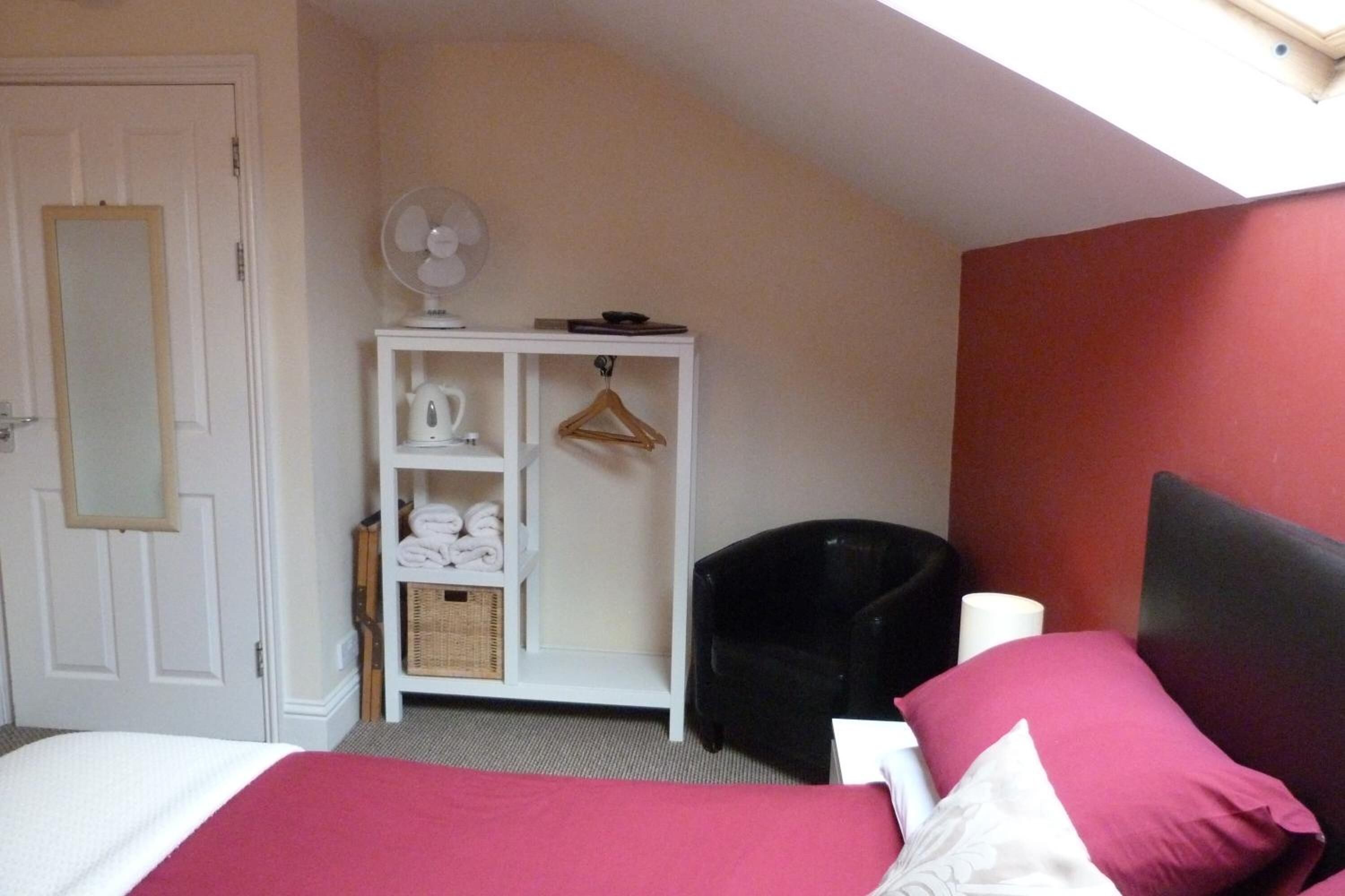 A bedroom with a bed, a white wardrobe, a black chair, a fan, and a red wall.