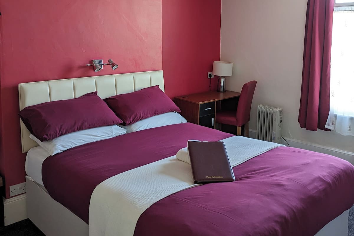 A hotel room with a large bed, a desk with a chair, a lamp, and a radiator.