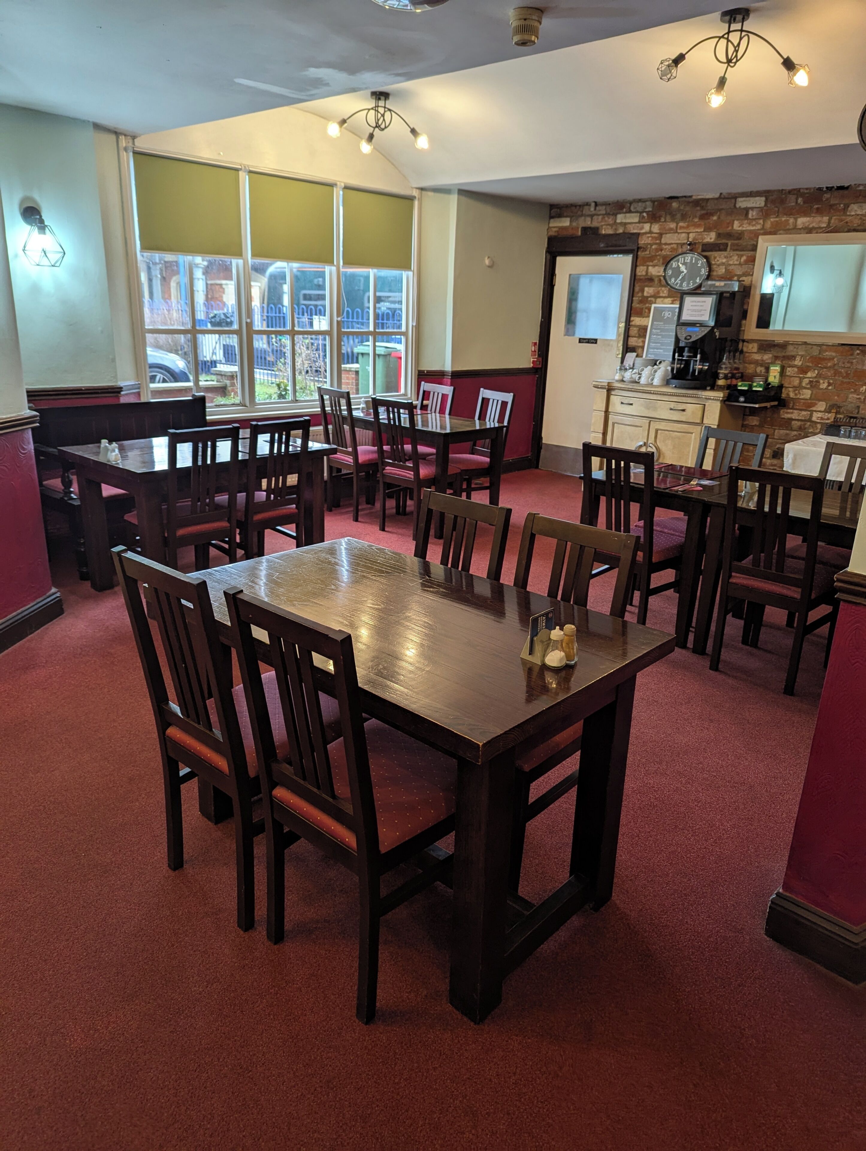 A dining area with wooden tables and chairs, a red carpet floor, and a brick wall with a clock.