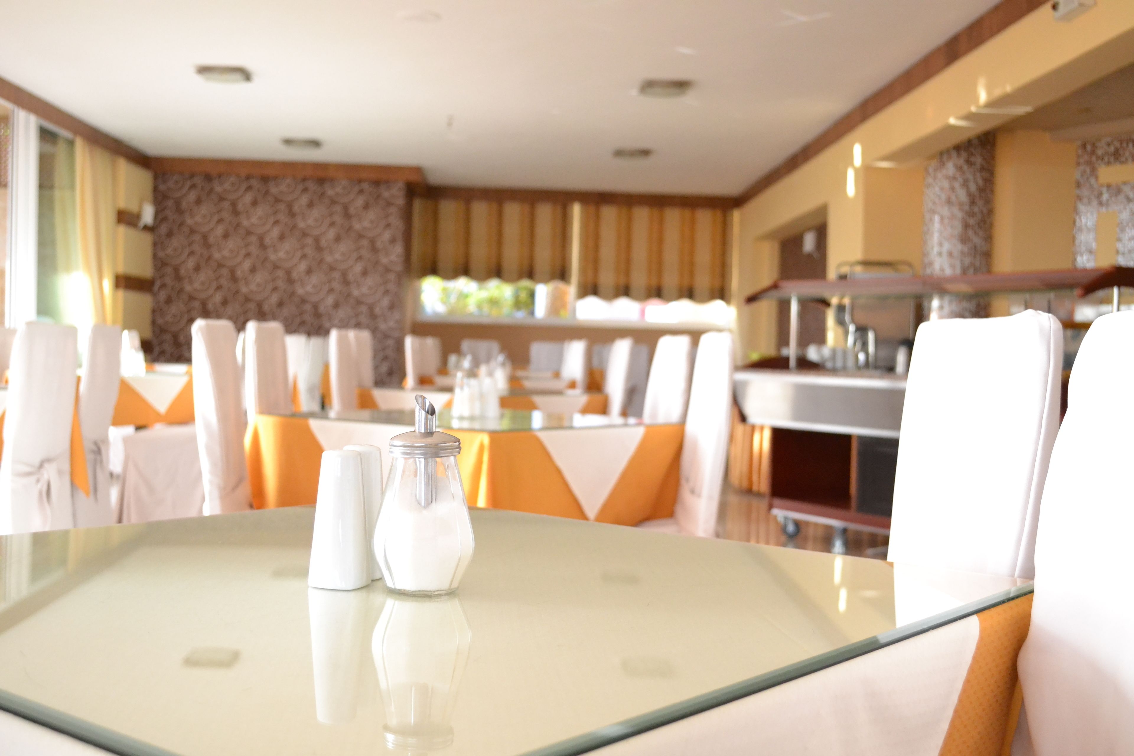 A dining area with tables set for service, a kitchen area in the background, and a patterned wall.