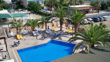 Piscina con tumbonas y palmeras, rodeada de edificios y vista al mar.