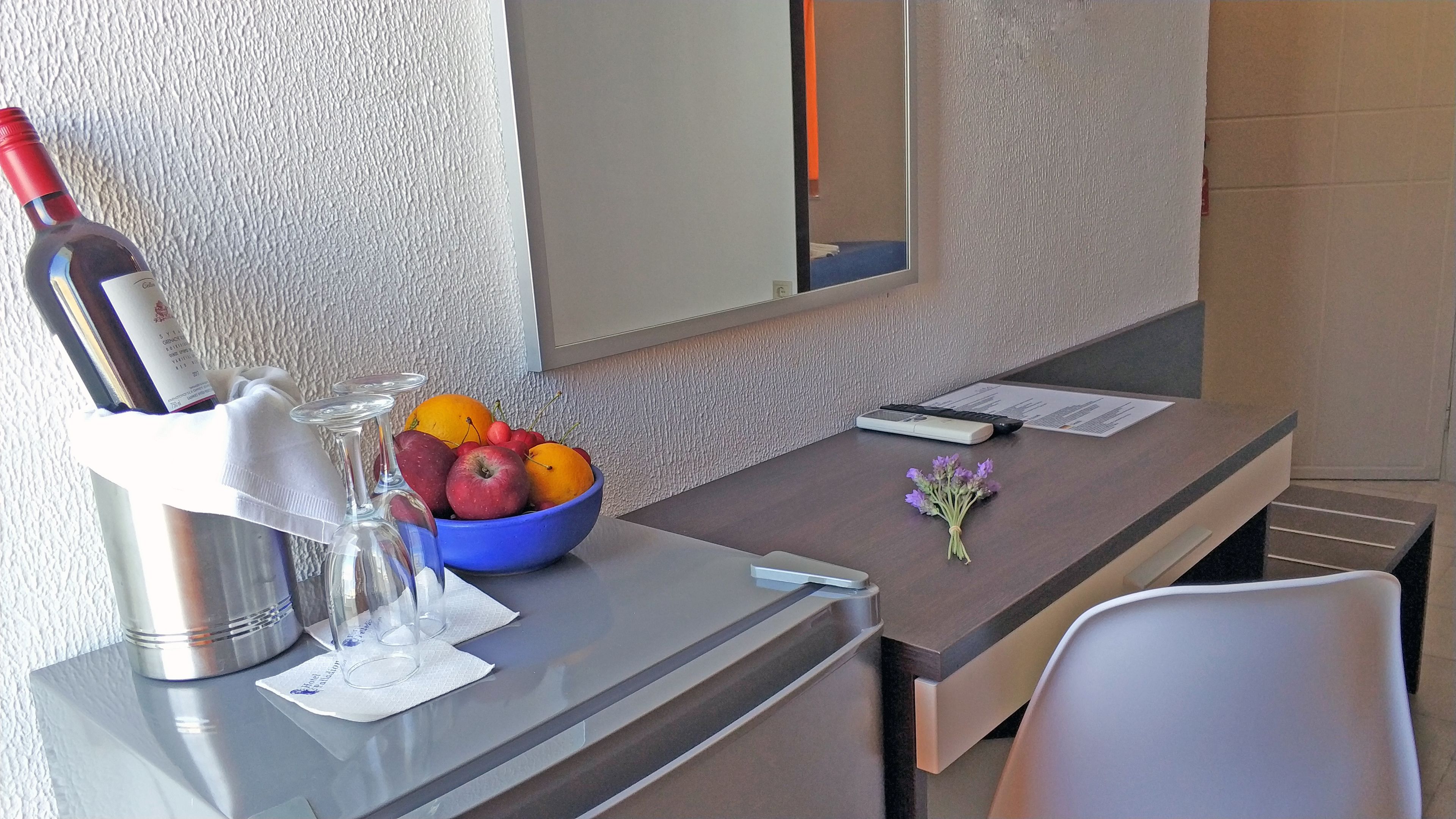 Escritorio de hotel con una botella de vino, copas, un frutero, un ramo de flores y un control remoto.