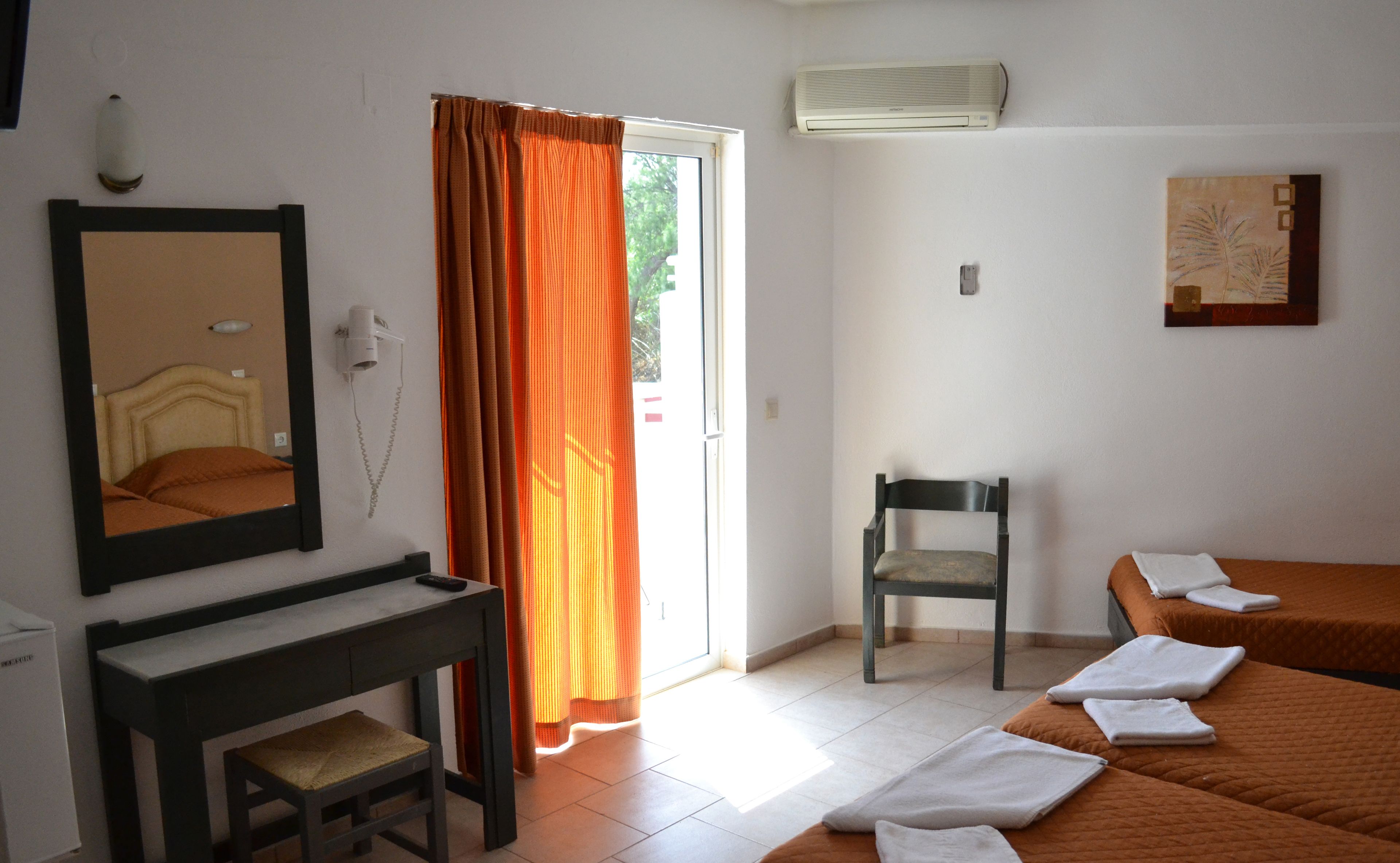 A hotel room with a bed, a chair, a desk, a mirror, an air conditioner, and a door with curtains.