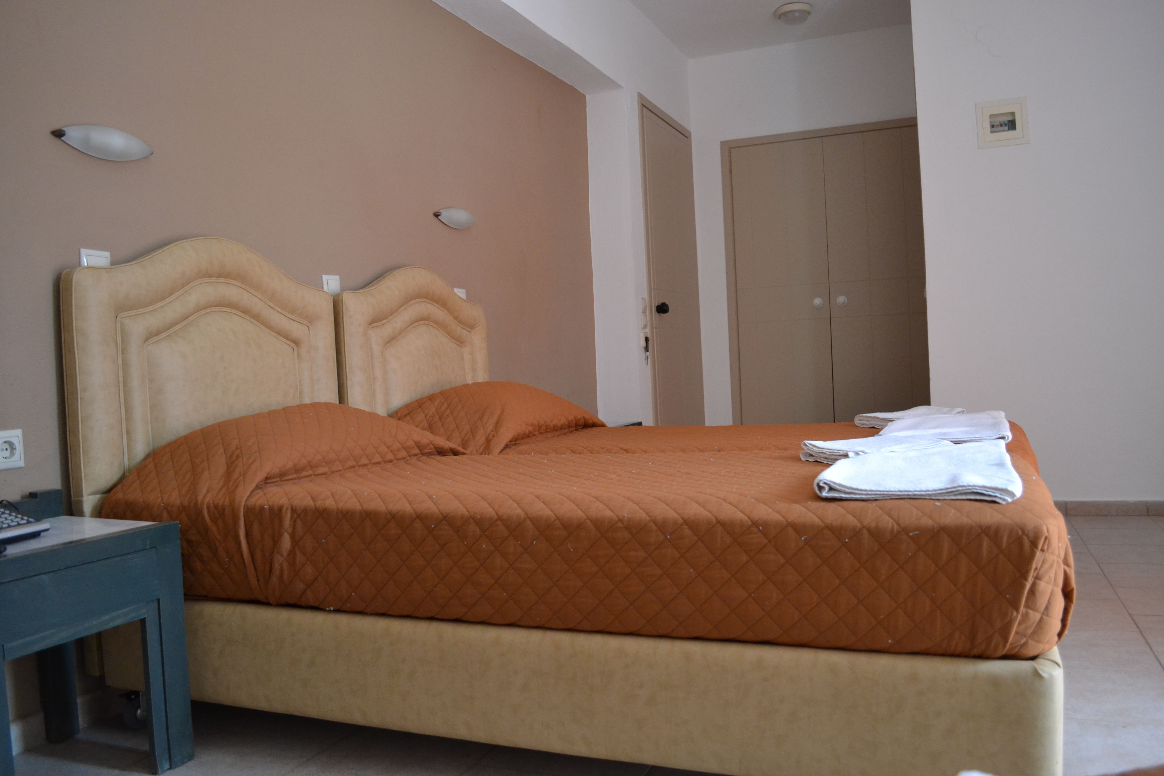 A neatly made bed with an orange quilt, a bedside table with a lamp, and a door in the background.