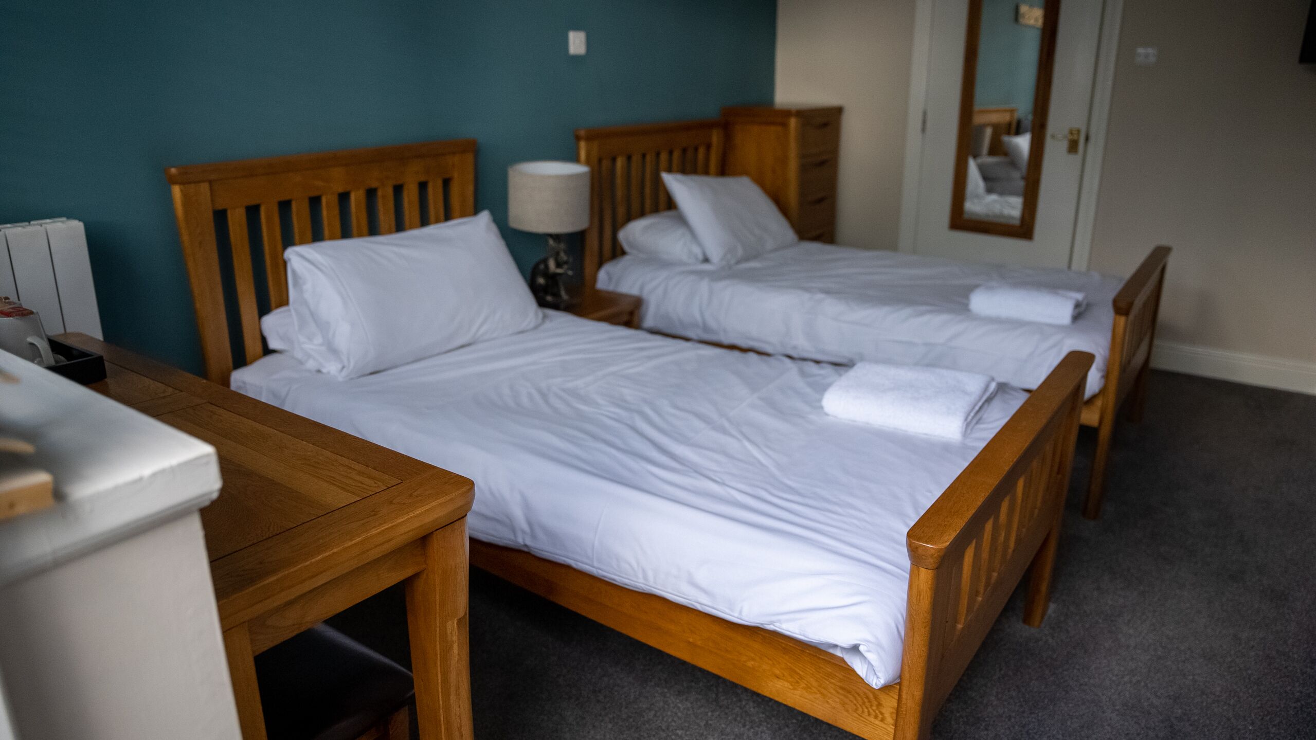 A hotel room with two wooden beds, a bedside table, a lamp, and a mirror.