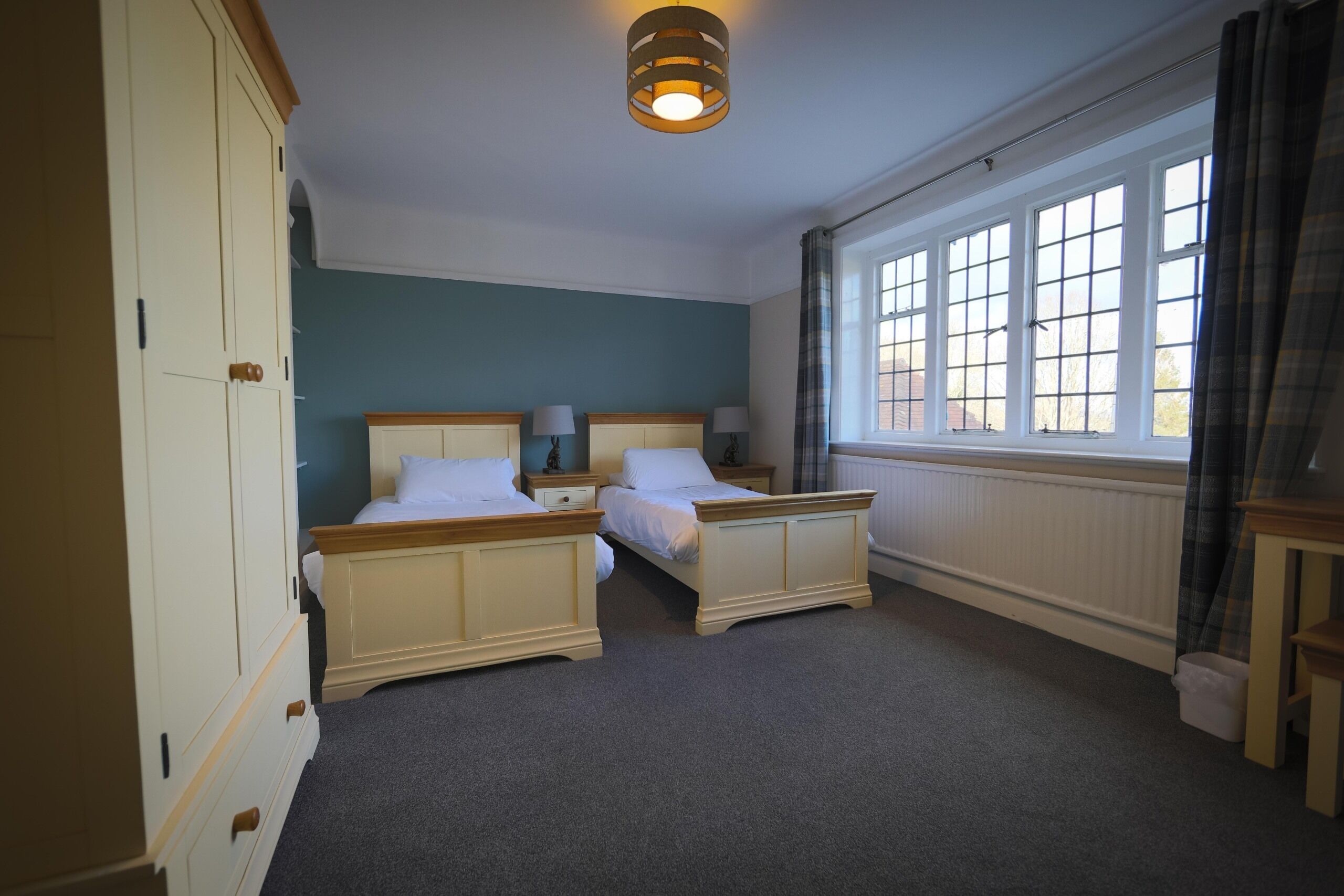 A bedroom with two single beds, a wardrobe, a window with curtains, and a ceiling light fixture.