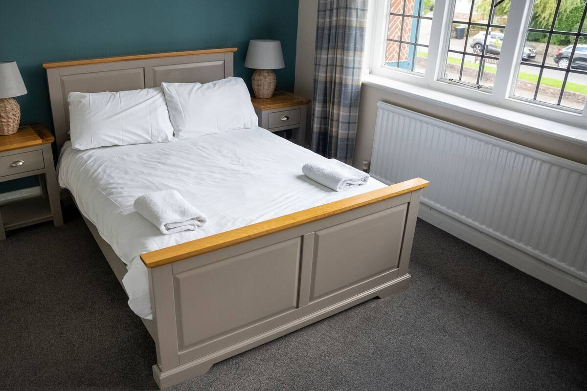 A neatly made bed with white linens and towels, a wooden headboard, two bedside tables with lamps, and a window with a view of the outside.