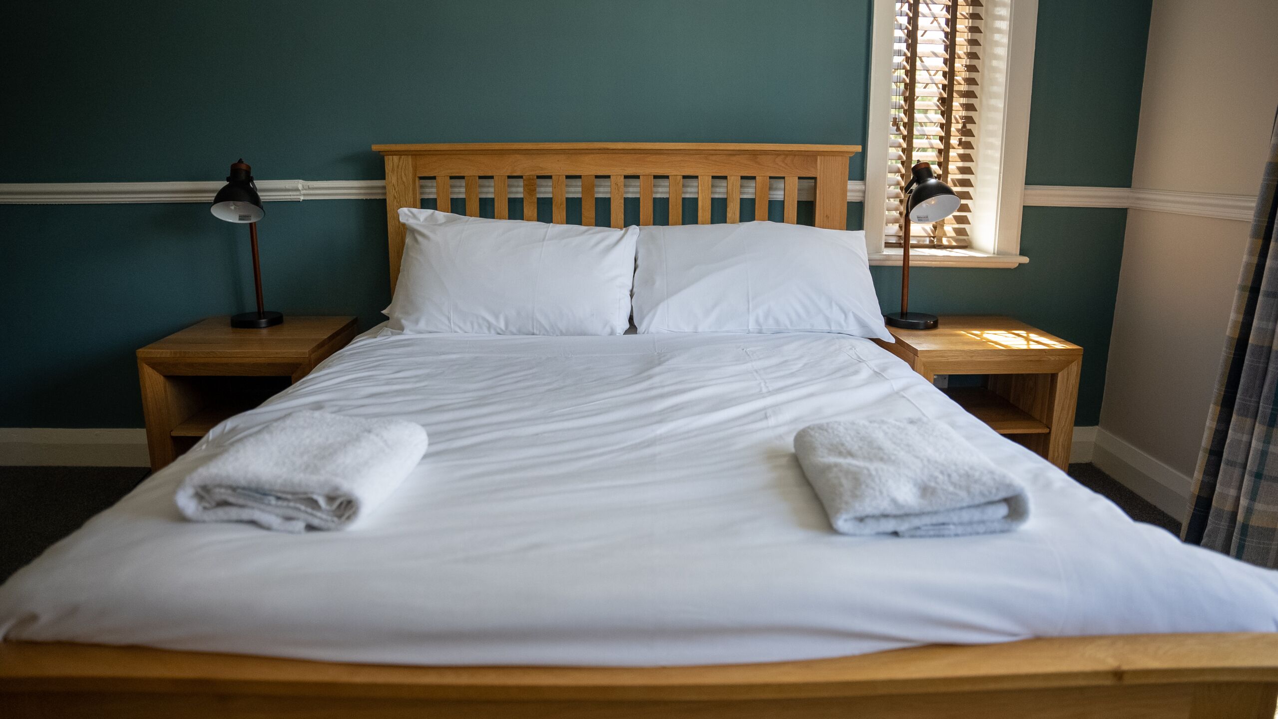 A neatly made bed with white linens and two towels folded at the foot. There are two bedside tables with lamps, and a window with blinds in the background.