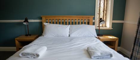 A neatly made bed with white linens and two towels folded at the foot. There are two bedside tables with lamps, and a window with blinds in the background.