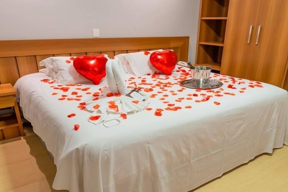 Un lit bien fait, recouvert d’une couvre-lit blanche, avec deux coussins en forme de cœur, un vase en verre et des pétales de rose éparpillés.