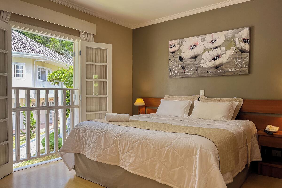 Une chambre avec un grand lit, une table de chevet avec une lampe, un tableau au mur et un balcon donnant sur l’extérieur.