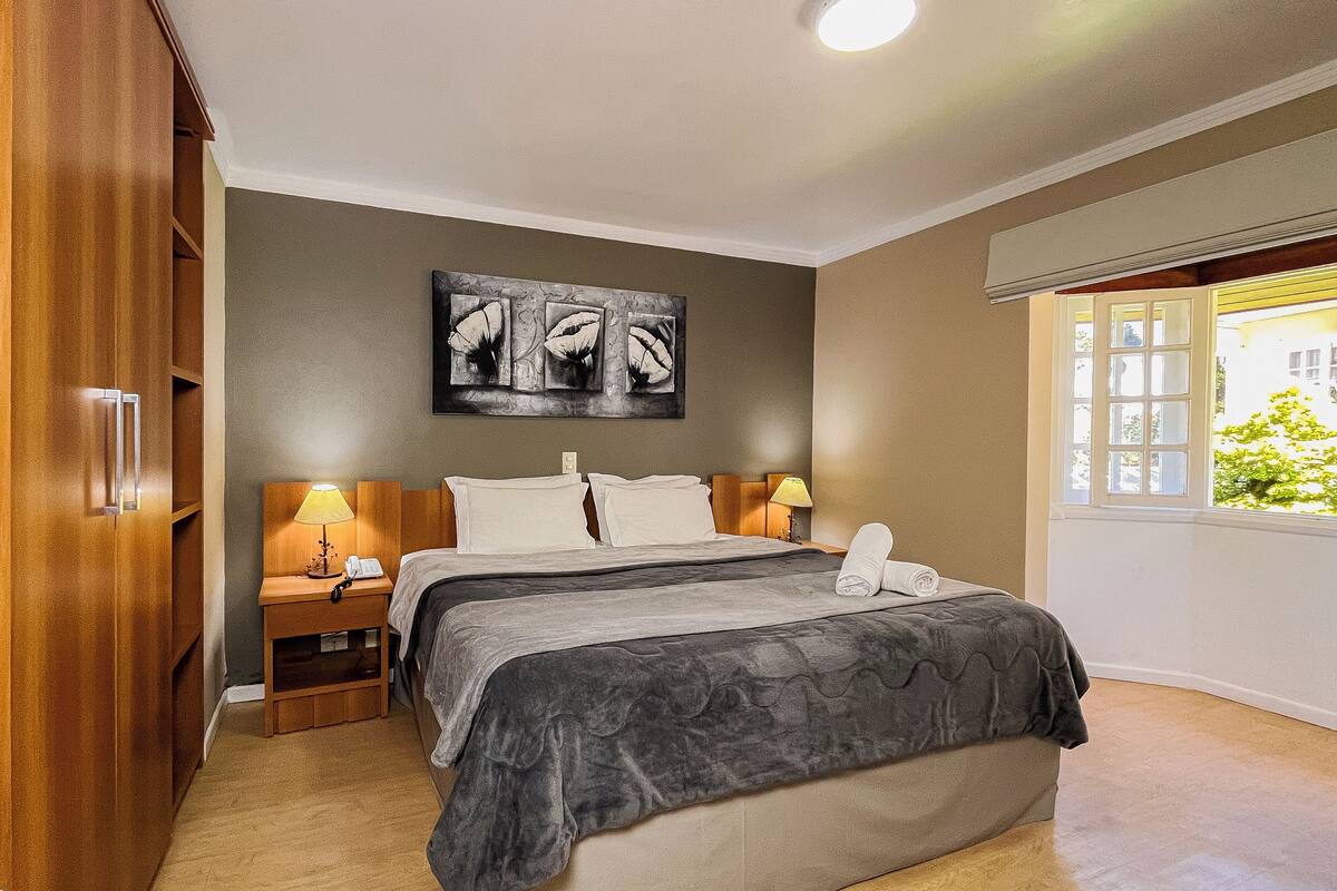 Une chambre à coucher comprenant un lit, des tables de chevet, une commode, une armoire, un tableau et une fenêtre donnant sur un paysage.