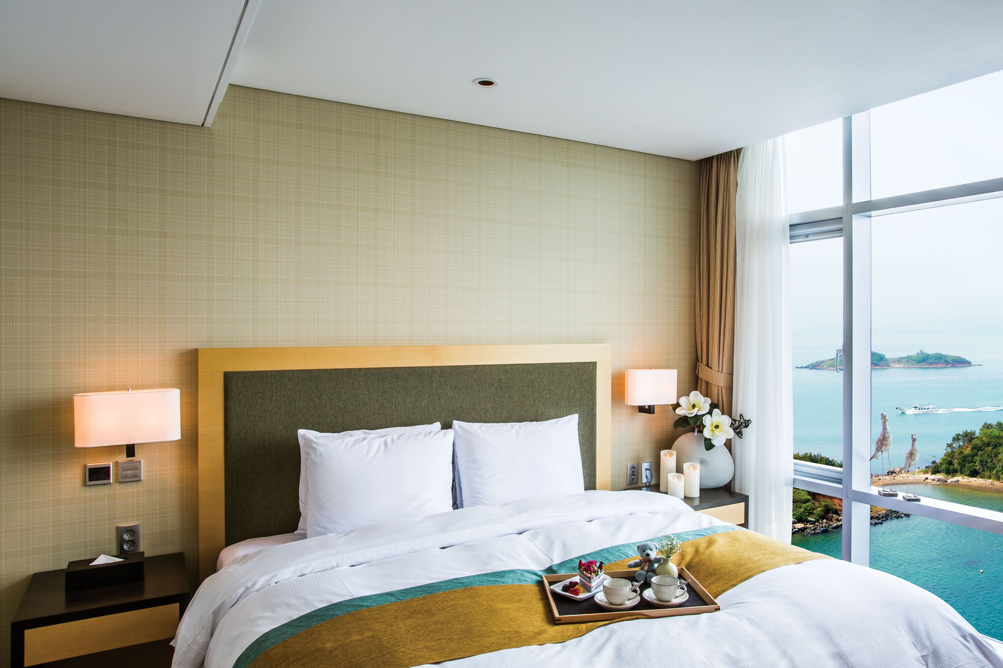 Habitación de hotel con una cama grande, dos lámparas de noche, una mesita de noche, una bandeja con alimentos para el desayuno y vistas al mar.