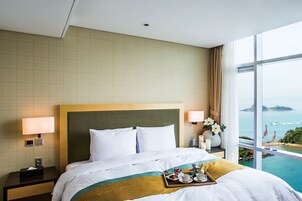 Habitación de hotel con una cama grande, dos lámparas de noche, una mesita de noche, una bandeja con alimentos para el desayuno y vistas al mar.