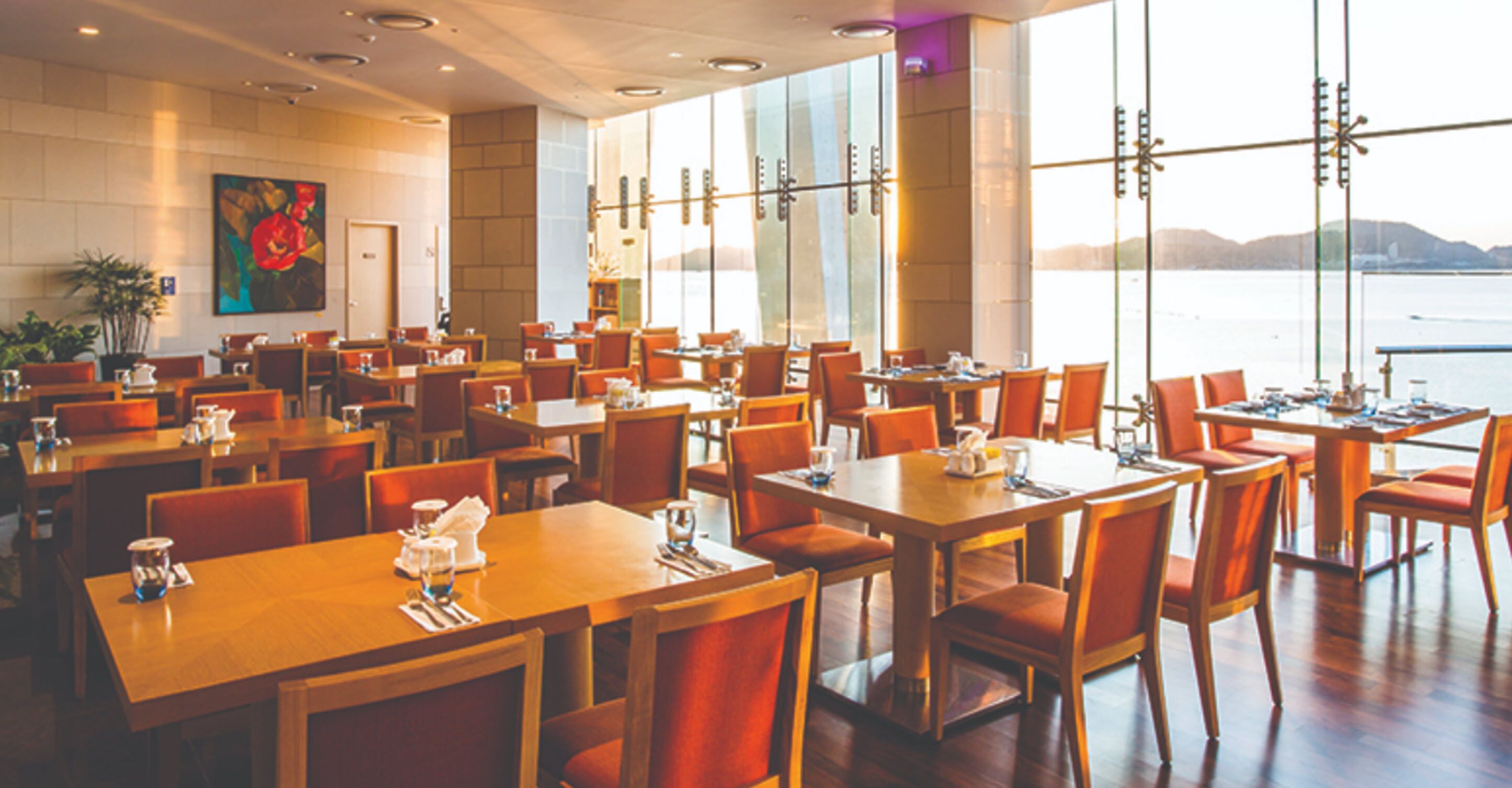 A spacious dining area with multiple tables and chairs, large windows offering a view of the water, and a painting on the wall.