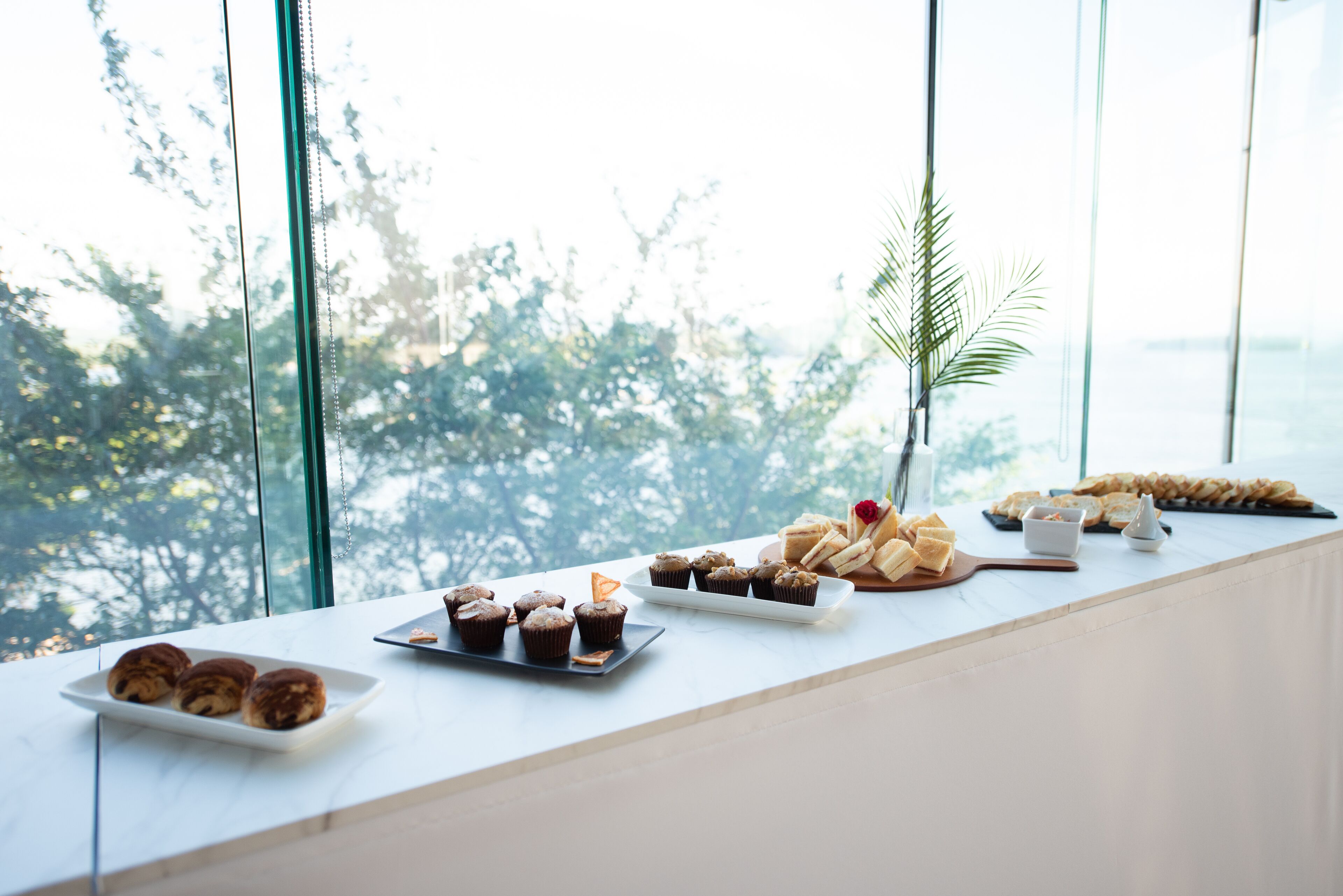 Een ontbijtbuffet met gebak en muffins op een witte tafel bij een groot raam met uitzicht op bomen.
