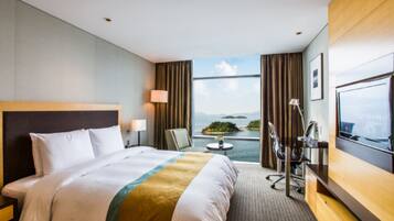 A hotel room with a large bed, a desk, a chair, a TV, and a view of the sea.