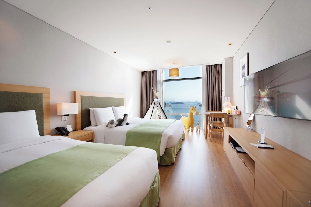 Quarto de hotel com duas camas, uma escrivaninha, uma cadeira, televisão e janela com vista.