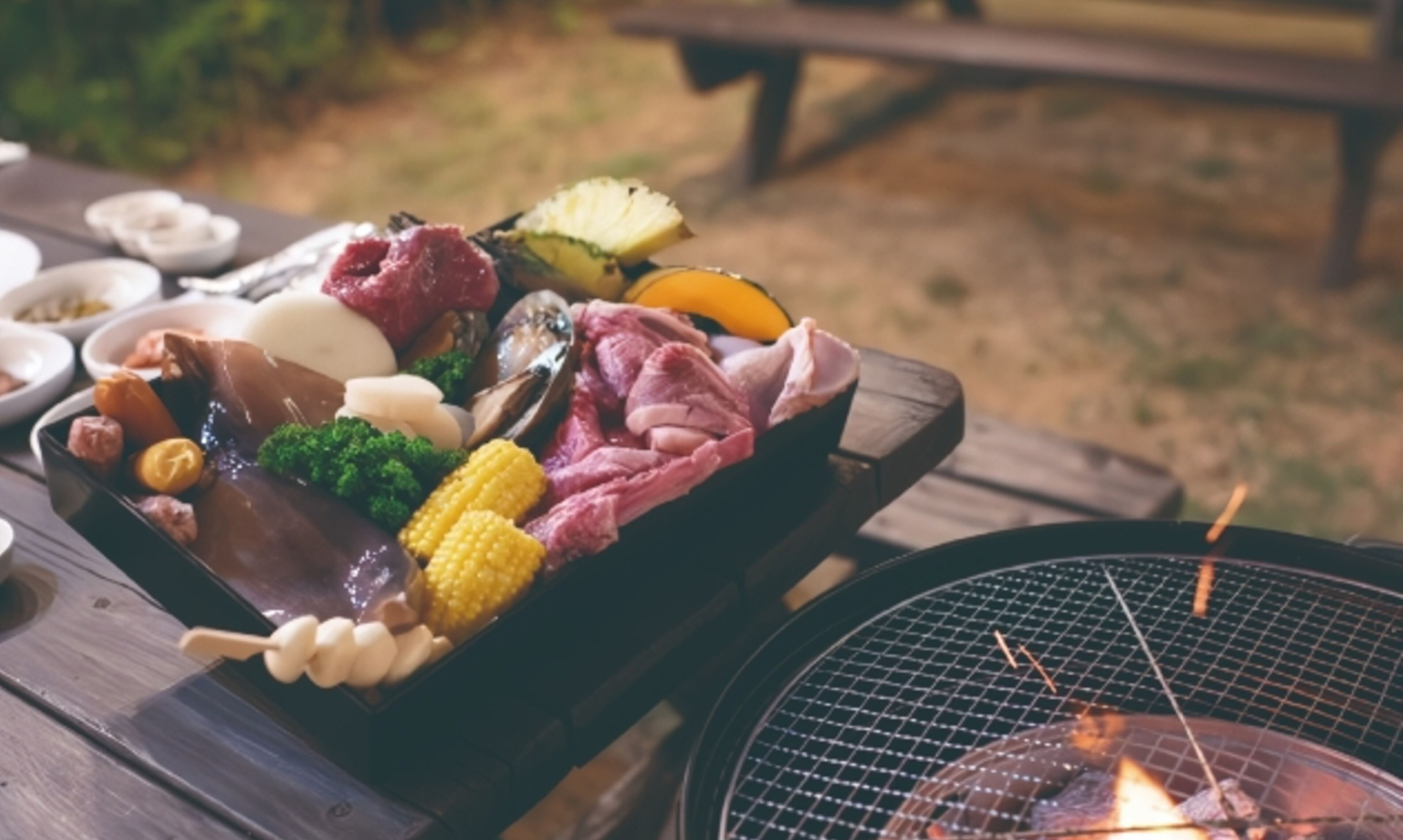 Una instalación para barbacoa con diferentes carnes y verduras sobre una mesa de madera, una parrilla con llamas visibles, y una mesa de picnic al fondo.