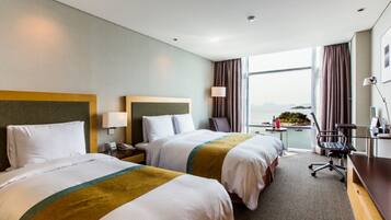 A hotel room with two beds, a desk, a chair, a lamp, and a view of the sea.