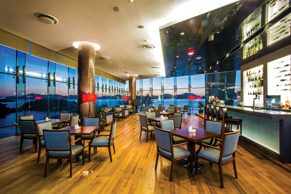 A restaurant with a view of the cityscape at dusk, featuring wooden tables and chairs, a bar area, and a large pillar.
