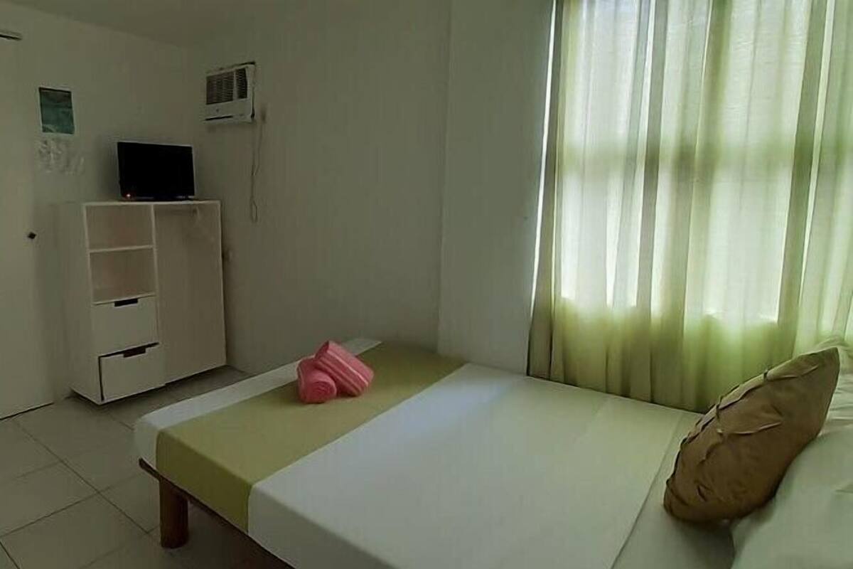 Ein kleiner, schlichter Raum mit einem Bett, einem Kleiderschrank, einem Fernseher und einem Fenster mit Vorhängen.