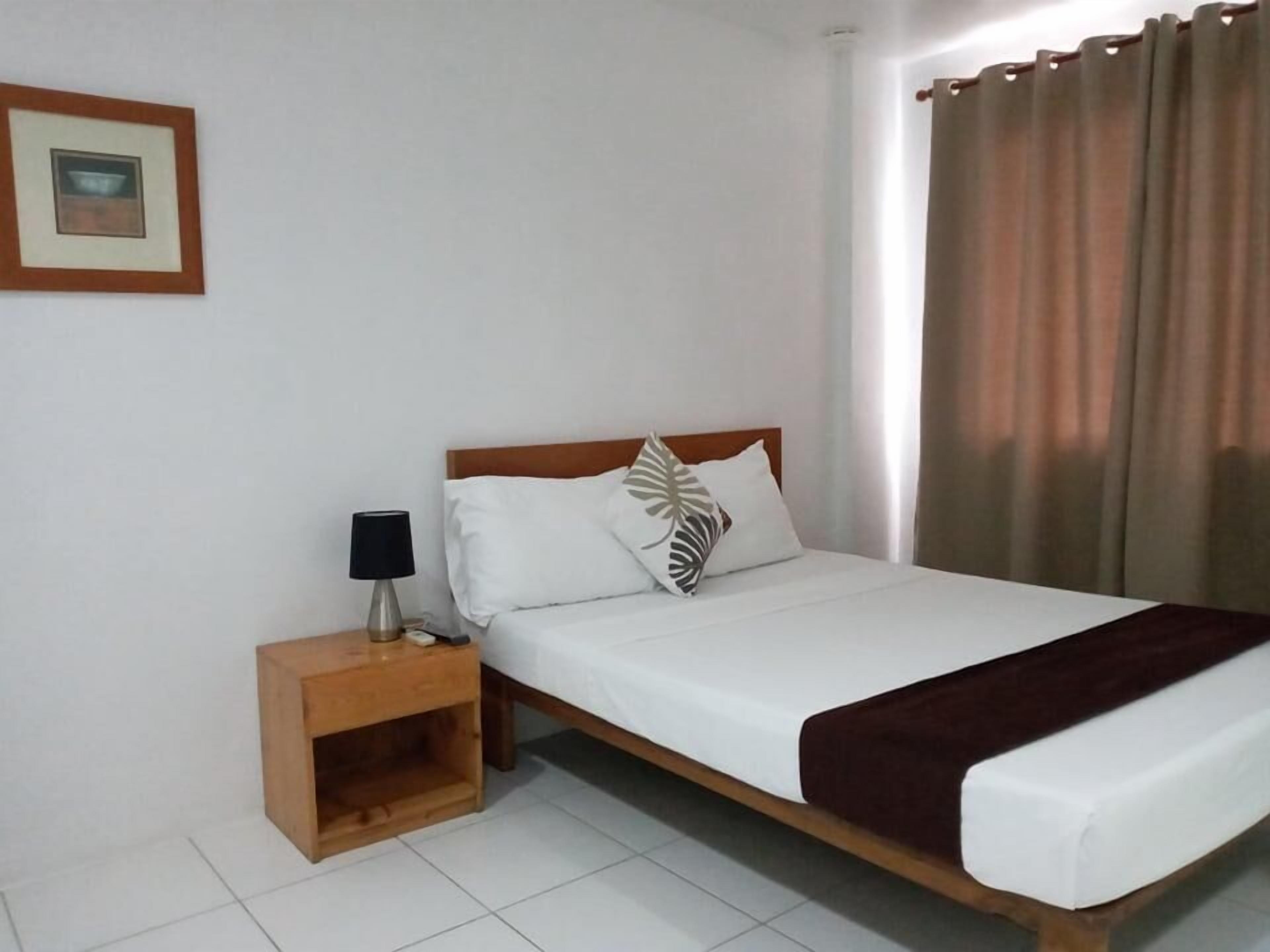 A single bed with a wooden headboard, white bedding, and a brown throw blanket. There is a nightstand with a lamp and a framed picture on the wall.
