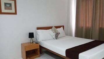 A single bed with a wooden headboard, white bedding, and a brown throw blanket. There is a nightstand with a lamp and a framed picture on the wall.