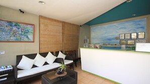 Een receptiegedeelte met een receptiebalie, een bank met witte kussens, een bijzettafeltje met een plant en een schilderij aan de muur.