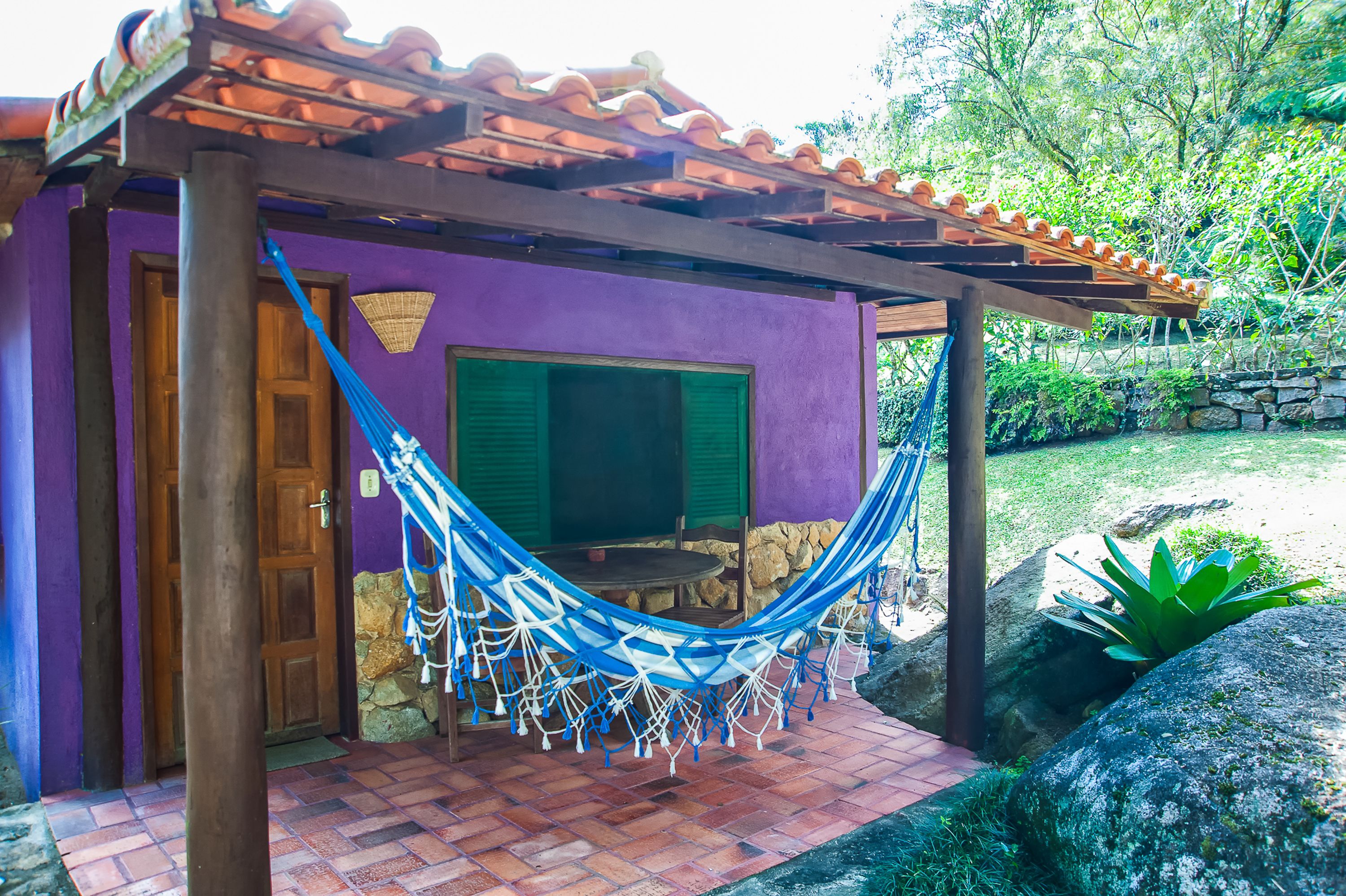 Un petit bâtiment d’un seul étage, à la façade violette, avec une porte en bois et un hamac suspendu sous une pergola en bois.