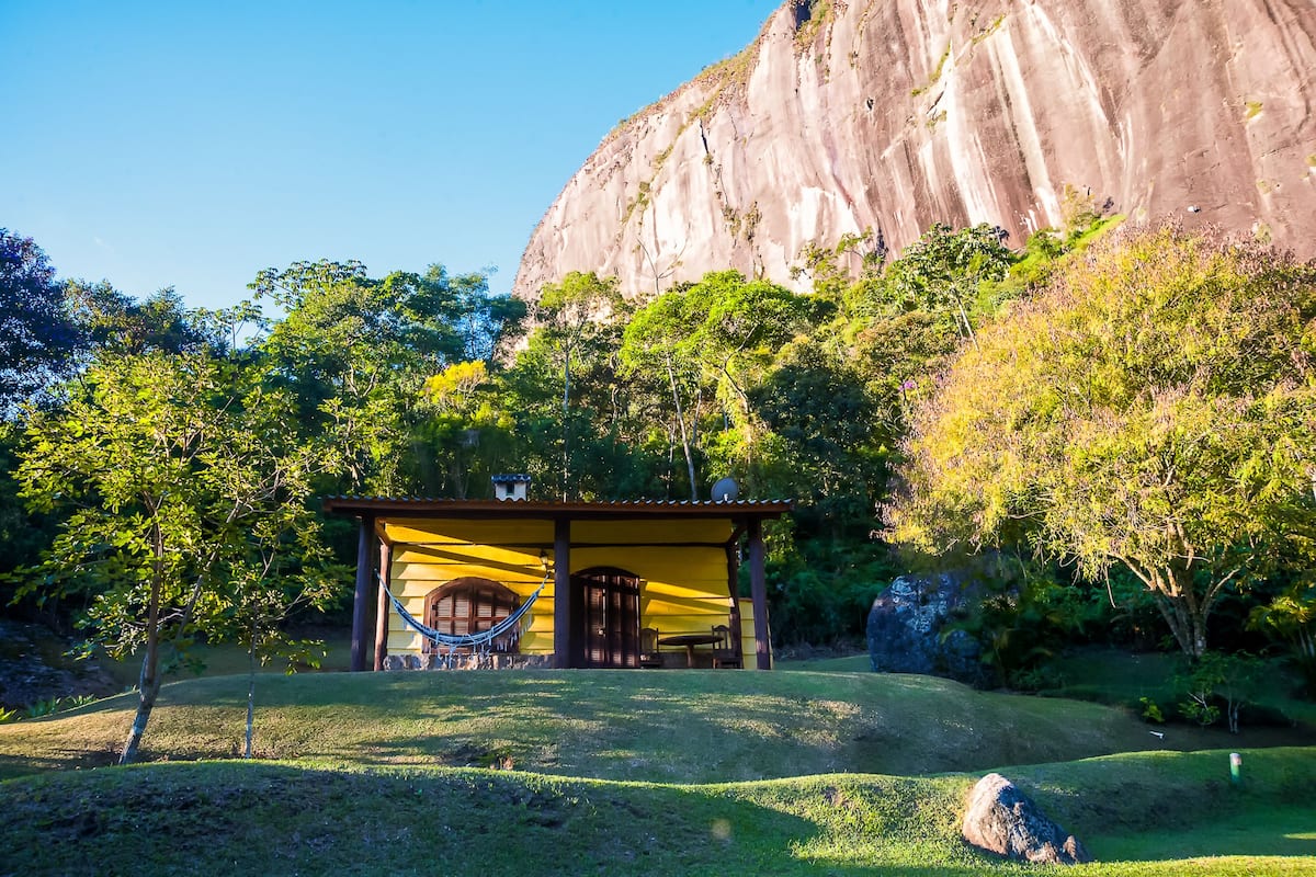 A small yellow building with a hammock and a table in front, surrounded by lush greenery and a large rock formation in the background.