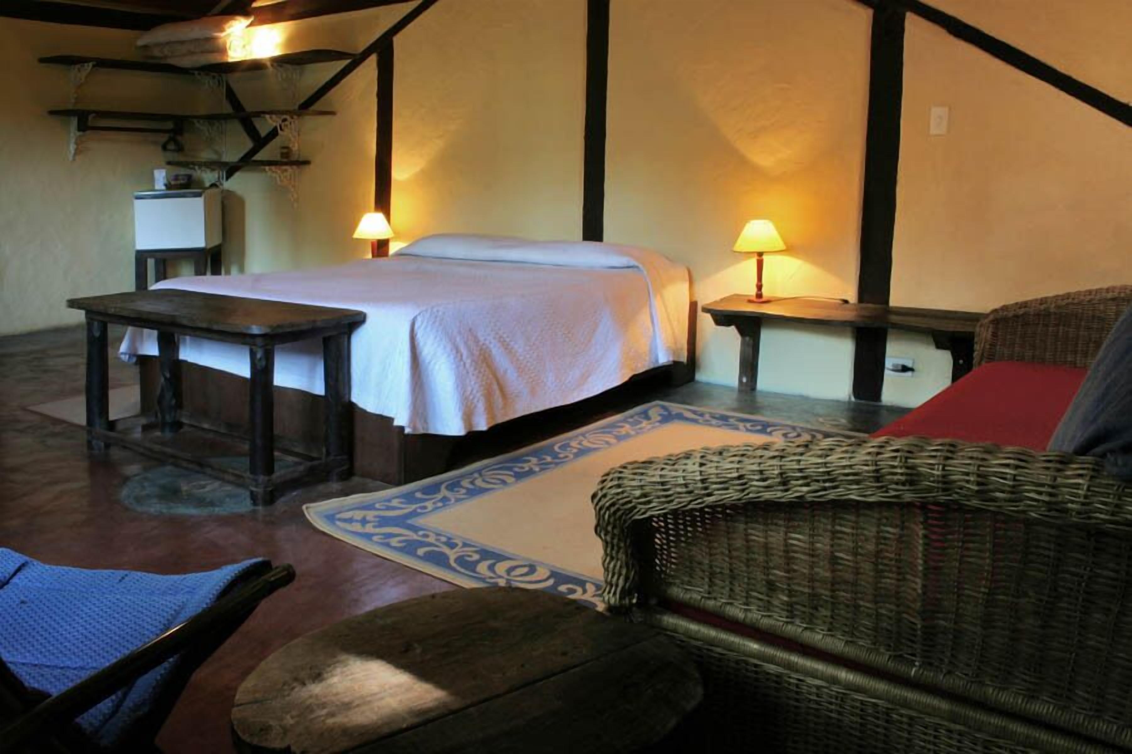 A room with a bed, a table, a chair, and a sofa. There are two lamps on the walls.